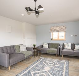A living room with three grey sofas a patterned rug and a window with a shade at Rynys in Llanddeiniolen near Llanrug