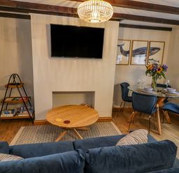 A living room with a sofa a round wooden coffee table a glass dining table with blue chairs and a wall mounted TV at April Cottage in Whitby