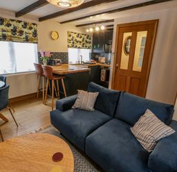 A living area with a blue sofa, a dining table with chairs, a kitchen island with bar stools, windows with patterned blinds, and a wooden door at April Cottage in Whitby