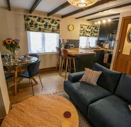 A living area with a blue sofa, round wooden coffee table, dining table with chairs and flowers, kitchen with bar stools and wooden door at April Cottage in Whitby