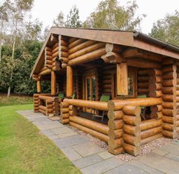 A wooden log cabin with a covered porch and green chairs surrounded by trees at Brynallt Country Park in Frankton near Ellesmere