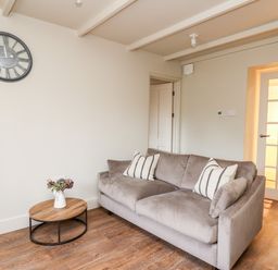 A living room with a gray sofa two striped pillows a round wooden coffee table with a vase of flowers and a wall clock at Llygad Y Dydd in Bow Street Wales