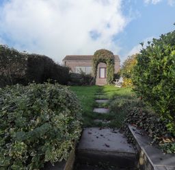 A stone path through a garden with bushes leading to a small building with a door covered by an arch of climbing plants at Hillcrest Oxwich near Port Eynon