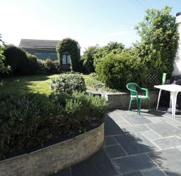 A garden with bushes lawn and a small building viewed from a stone patio with plastic chairs and a white table at Hillcrest in Oxwich near Port Eynon