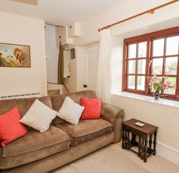 A living room with a brown sofa and cushions next to a window with a vase of flowers on the sill at Hillcrest Oxwich near Port Eynon