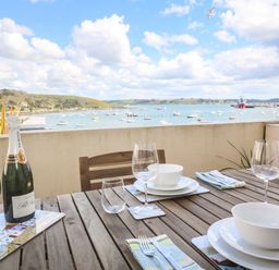 A wooden outdoor dining table set with glassware, plates, bowls, and a bottle of champagne overlooking a harbor with boats at 58 High Street in Falmouth