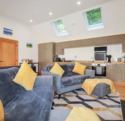 A living room with two grey sofas with yellow cushions and a geometric rug next to a kitchen with brown cabinets and skylight windows at Clonmore in Bodmin