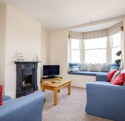 A living room with blue sofas red cushions a wooden coffee table and a window seat at Driftwood in Fowey