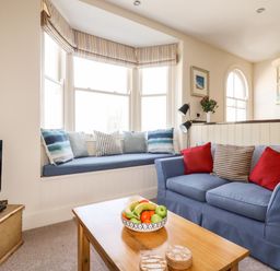 A sitting area with a blue sofa and striped cushions next to a window seat with cushions and a wooden coffee table with a bowl of fruit at Driftwood in Fowey