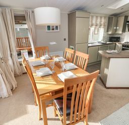A dining area with a wooden table and six chairs set with white dishes next to a kitchen with gray cabinets and a window at Hafan Lodge 85 Wisemans Bridge near Saundersfoot