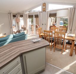 An open plan living and dining area with a kitchen counter a blue sofa a wooden dining table with six chairs and large windows at Hafan Lodge 85 Wisemans Bridge near Saundersfoot