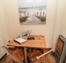 A small dining area with a wooden table and two chairs a folding chair leaning against the wall and a beach painting on the wall at Coras House in Amble
