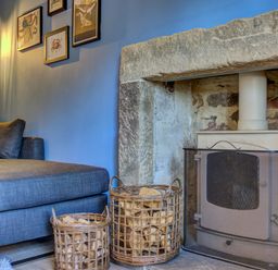 A living area with a gray sofa next to a stone fireplace with a wood stove and wicker baskets filled with firewood at Crab Cottage in Sandsend