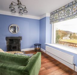 A sitting room with a green sofa a small round table a fireplace and a large window with a floral curtain at Estcote Roger Ground near Hawkshead