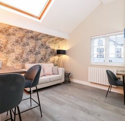 A living area with a sofa, patterned wall, floor lamp, dining table with chairs, and a window with blinds at Fentys in Bowness-On-Windermere