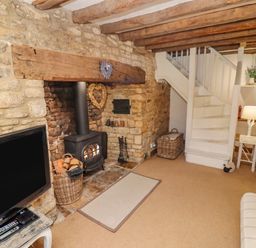 A living room with a wood-burning stove in a stone fireplace a television on a small table a white staircase and striped sofas at Chippy Cottage in Chipping Norton