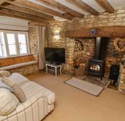 A living room with wooden beams stone walls a sofa a window seat a television and a fireplace at Chippy Cottage in Chipping Norton