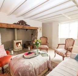 A living room with a fireplace armchairs a sofa a covered ottoman and flowers on a tray at Holly Cottage in Minster Lovell