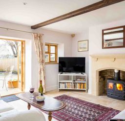 A living room with a wood burning stove TV on a white stand and an open door to a garden at Oyster Barn in Naunton