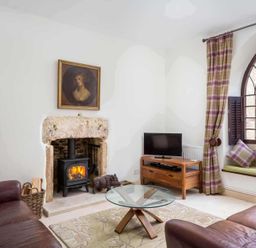 A living room with a fireplace a wood stove two leather sofas a glass coffee table a television on a wooden stand and a window seat with cushions at Little Culver in Minchinhampton