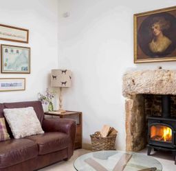 A living room with a brown leather sofa pillows a wood burning stove in a stone fireplace and framed pictures on the wall at Little Culver in Minchinhampton