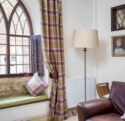 A seating area with a window seat with cushions and plaid curtains next to a floor lamp and a leather sofa at Little Culver in Minchinhampton