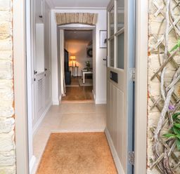 Open front door leading to hallway with built-in white cabinets and view into living area with lamp and table at Spinners Cottage in Stow-on-the-Wold