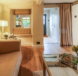 A sitting area with a beige sofa and side table with a lamp and decorative items and a window seat next to a passage with a curtain and a glass coffee table with magazines and flowers at Spinners Cottage in Stow-on-the-Wold
