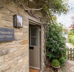 The entrance of a stone cottage with an open door a wall sign reading Spinners Cottage plants and a small wooden gate at Spinners Cottage in Stow-on-the-Wold