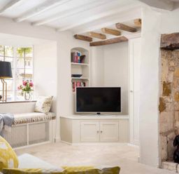 A living room with a window seat flowers on the window ledge a television built into a white cabinet exposed wooden ceiling beams and a stone fireplace at Stanley Cottage in Chipping Campden