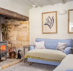 A living room with a wood burning stove stone wall and two blue sofas at Stanley Cottage in Chipping Campden