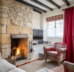 A living room with a stone fireplace a TV an armchair and red curtains at Lanes Cottage in Chipping Campden