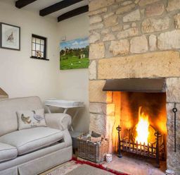 A living room with a stone fireplace with a fire a beige sofa and a small white table at Lanes Cottage in Chipping Campden