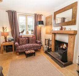 A living room with a fireplace a patterned loveseat a floor lamp a side table with a lamp and books and a television on a stand at Green Pump Cottage in Blockley