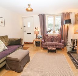 A living room with a green sofa an ottoman a purple patterned armchair a wooden side table and a fireplace at Green Pump Cottage in Blockley