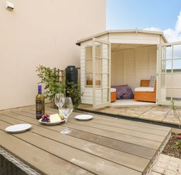 An outdoor patio with a wooden table set with wine glasses and a bottle near a white garden shed with open doors and wicker chairs inside at Pebbles Reach in Fortuneswell