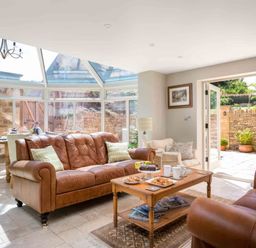 A sunroom with leather sofas, a wooden coffee table with snacks and drinks, dining table and chairs visible through glass walls at East Leaze in Chipping Campden