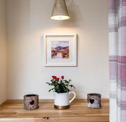 A wooden countertop with two heart cutout candle holders and a white jug with red flowers under a wall light and framed landscape painting at Eastnor in Moreton-in-Marsh