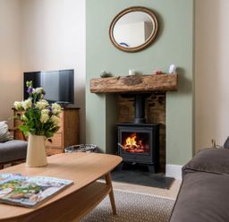 A living room with a wood burning stove under a wooden beam mantel a round mirror above and a television on a wooden cabinet in Wall Cottage in Burford