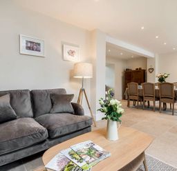 A living room with a gray sofa a wooden coffee table with flowers and magazines and a dining area with a table and six chairs at Wall Cottage in Burford