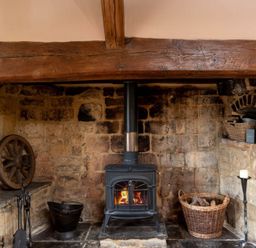 A stone fireplace with a wood burning stove a basket of firewood and fireplace tools at Little Gables in Broadway