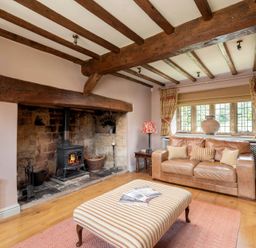 A living room with a wood burning stove stone fireplace leather sofas striped ottoman and wooden beams on the ceiling at Little Gables Broadway