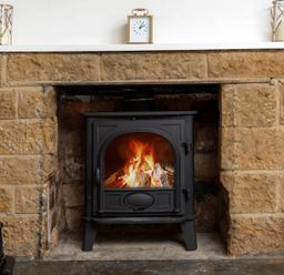 A lit wood burning stove in a stone fireplace with a clock on the mantel at Holly Cottage Middle Tysoe in Tysoe