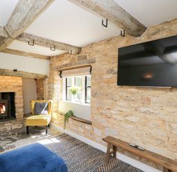 A living room with a stone fireplace yellow chair window with flowers blue ottoman and wall mounted television at Millbank Cottage in Mickleton
