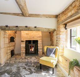 A room with a stone fireplace a yellow chair with a cushion and flowers on the window sill at Millbank Cottage in Mickleton