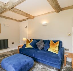 A seating area with a blue sofa and ottoman with yellow and patterned cushions in a room with stone flooring and wooden beams at Millbank Cottage in Mickleton
