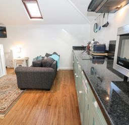 A kitchen with green cabinets a countertop and a microwave next to a living area with a gray sofa a side table and a wall mounted TV at The Bath House in Blockley