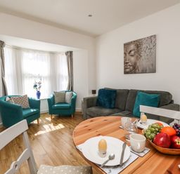 A living room with two teal armchairs, a grey sofa with pillows, a round wooden dining table set with plates and fruit bowl at Marina Reach in Weymouth