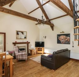 A living room with a wooden dining table chairs a plaid armchair a brown leather sofa a television a fireplace wooden beams and a spiral staircase at The Old Dairy in Maenclochog