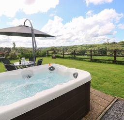 A hot tub in a garden with a table and chairs under an umbrella and a wooden fence with a view of trees at The Old Dairy in Maenclochog